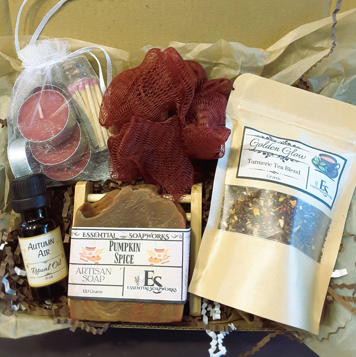 Autumn self-care ritual kit containing multiple wellness and spa items: a bottle of Autumn Air ritual oil, a bar of Essential Soapworks pumpkin spice soap, a pouch of Turmeric Tea Blend, a mesh red loofah, a candle in a white bag, and a small match pack, arranged in brown paper filler.