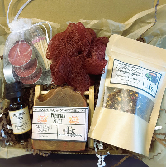 Autumn self-care ritual kit containing multiple wellness and spa items: a bottle of Autumn Air ritual oil, a bar of Essential Soapworks pumpkin spice soap, a pouch of Turmeric Tea Blend, a mesh red loofah, a candle in a white bag, and a small match pack, arranged in brown paper filler.