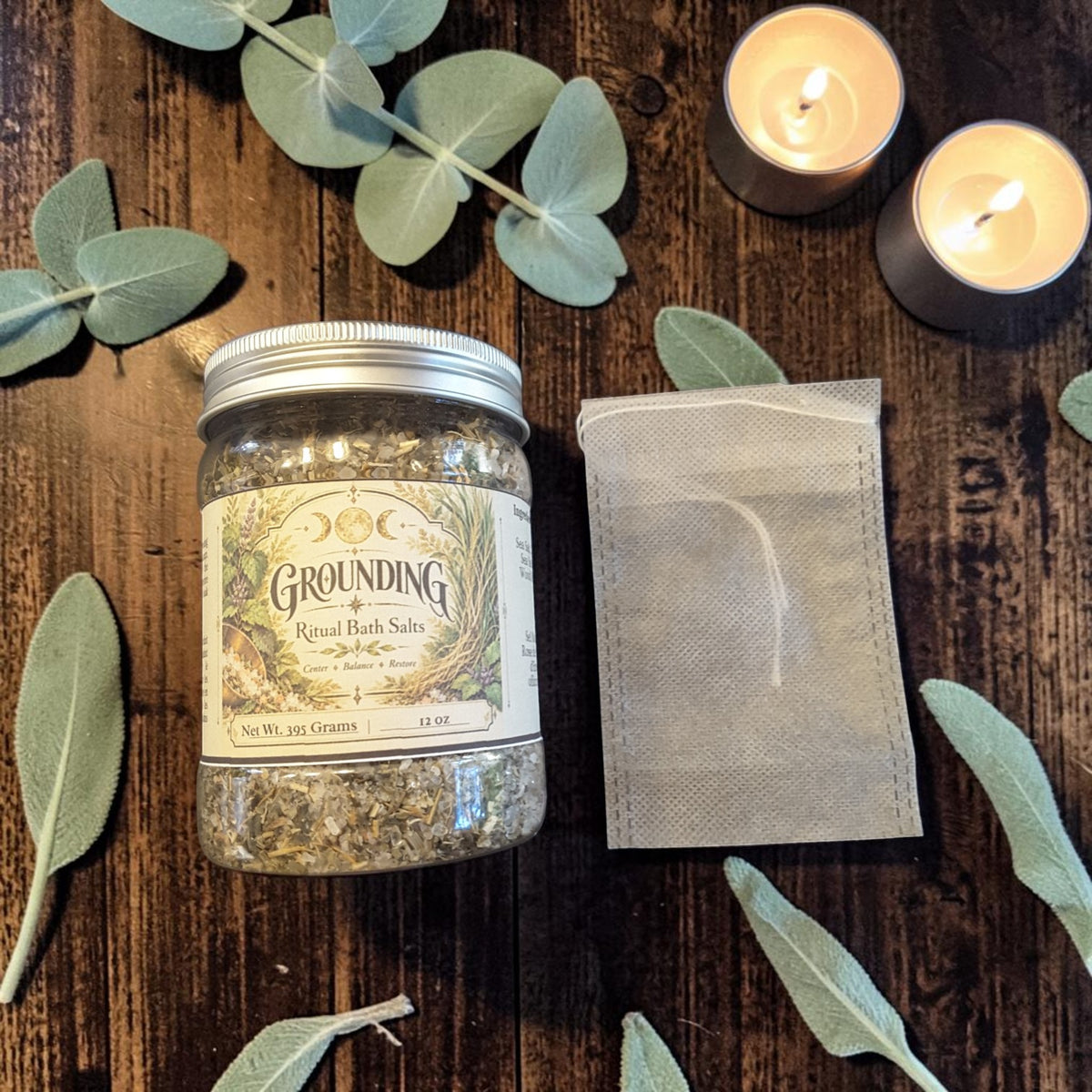 grounding bath salts in a jar alongside a muslin bag on a dark wooden surface with candles and greenery 