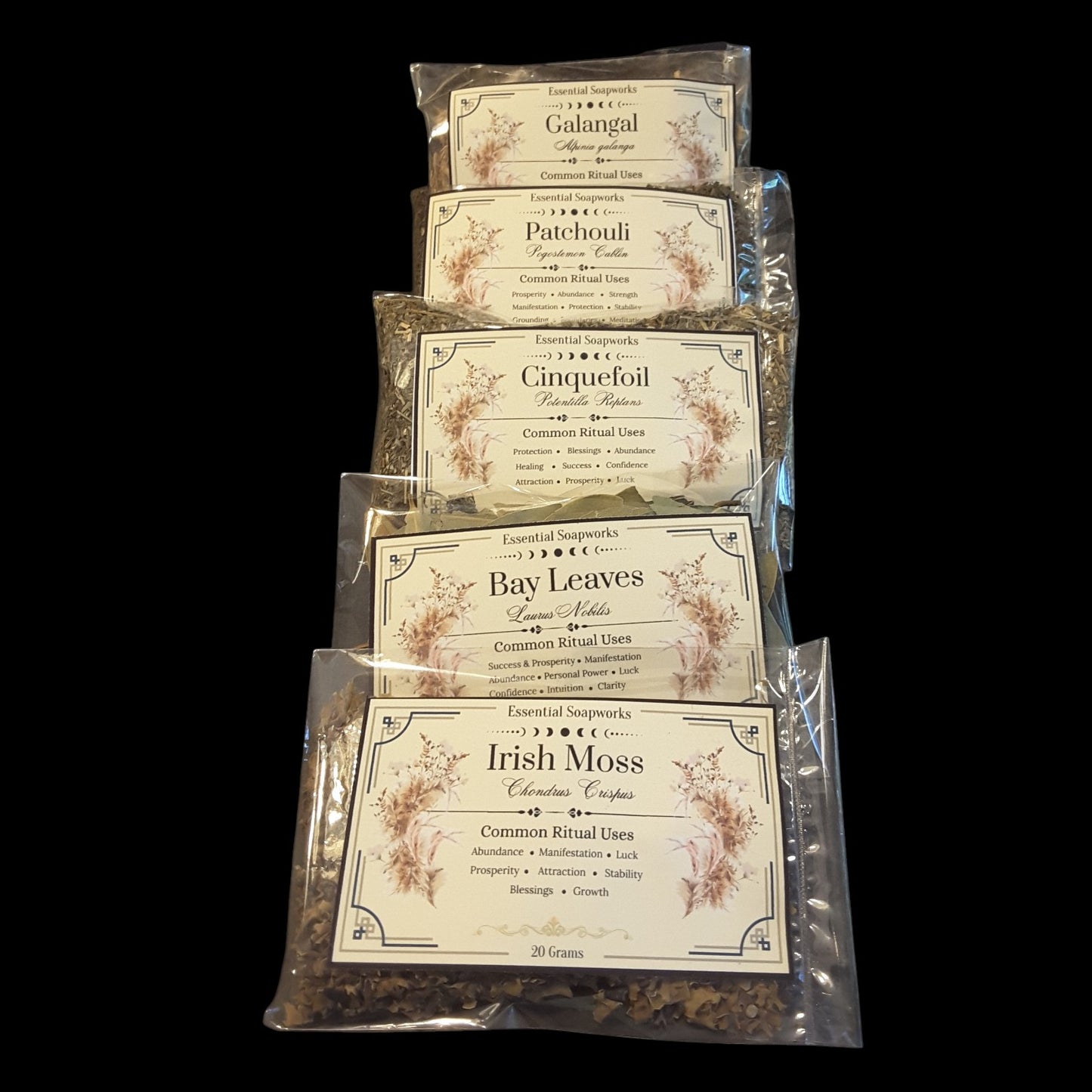 Six clear plastic bags of abundance and manifestation herbs for wealth and money magic arranged vertically on a black background. The herbs included are Galangal, Patchouli, Cinquefoil, Bay Leaves, and Irish Moss, with a top brand mark that appears to read Essential Soapworks.
