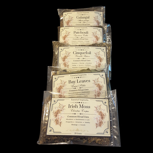 Six clear plastic bags of abundance and manifestation herbs for wealth and money magic arranged vertically on a black background. The herbs included are Galangal, Patchouli, Cinquefoil, Bay Leaves, and Irish Moss, with a top brand mark that appears to read Essential Soapworks.