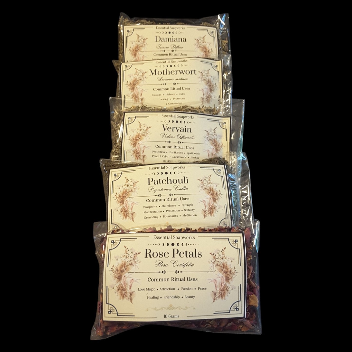 Five clear plastic bags of witchy herbs for love spells and rituals stacked vertically on a black background. The labels read Daminia, Motherwort, Vervain, Patchouli, and Rose Petals, with floral illustrations and common ritual uses printed on the cream-colored labels.