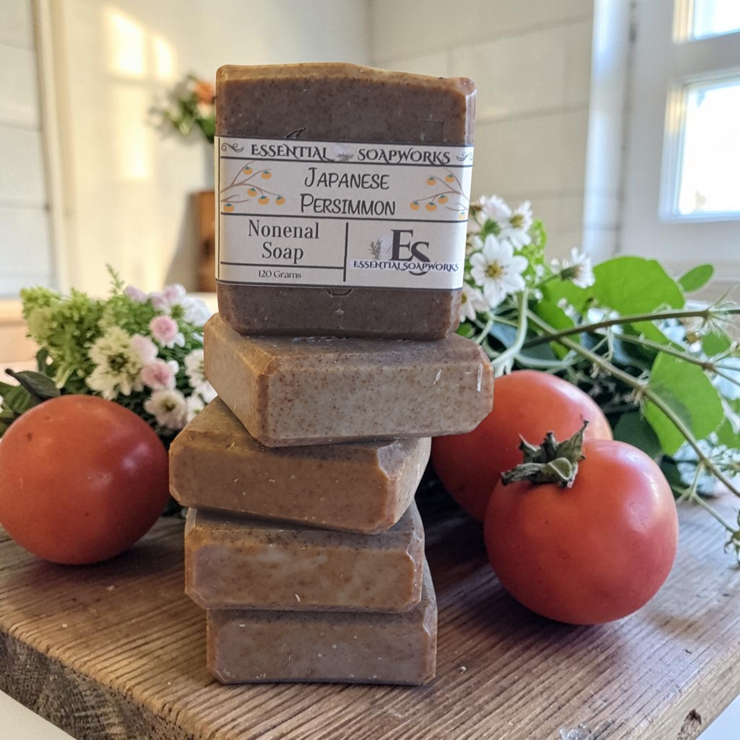 Stack of Persimmon Soap - Nonenal Soap bars with a visible label on a wooden surface with persimmons and flowers in the background