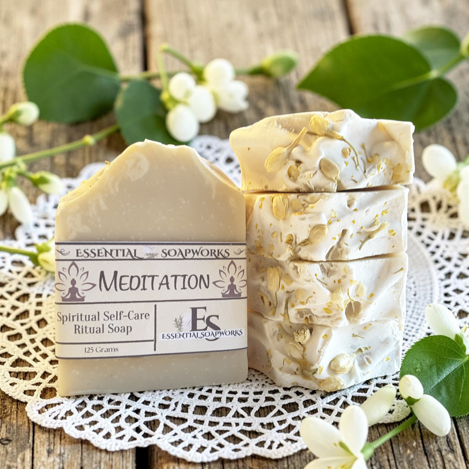 Two bars of Essential Soapsworks Meditation Spiritual Self-Care Ritual Soap displayed on a white lace doily over a rustic wooden surface, surrounded by white blossoms and green leaves.