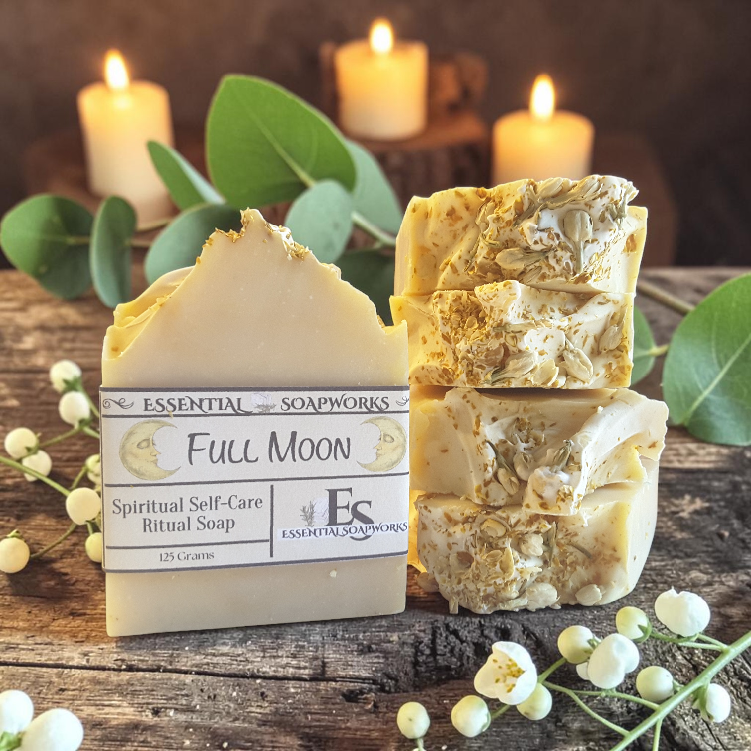 A bar of Essential Soapworks Full Moon Spiritual Self-Care Ritual Soap stands next to three stacked cut bars of the same handmade soap on a rustic wooden surface, with green leaves and white flowers around it and lit candles blurred in the background.