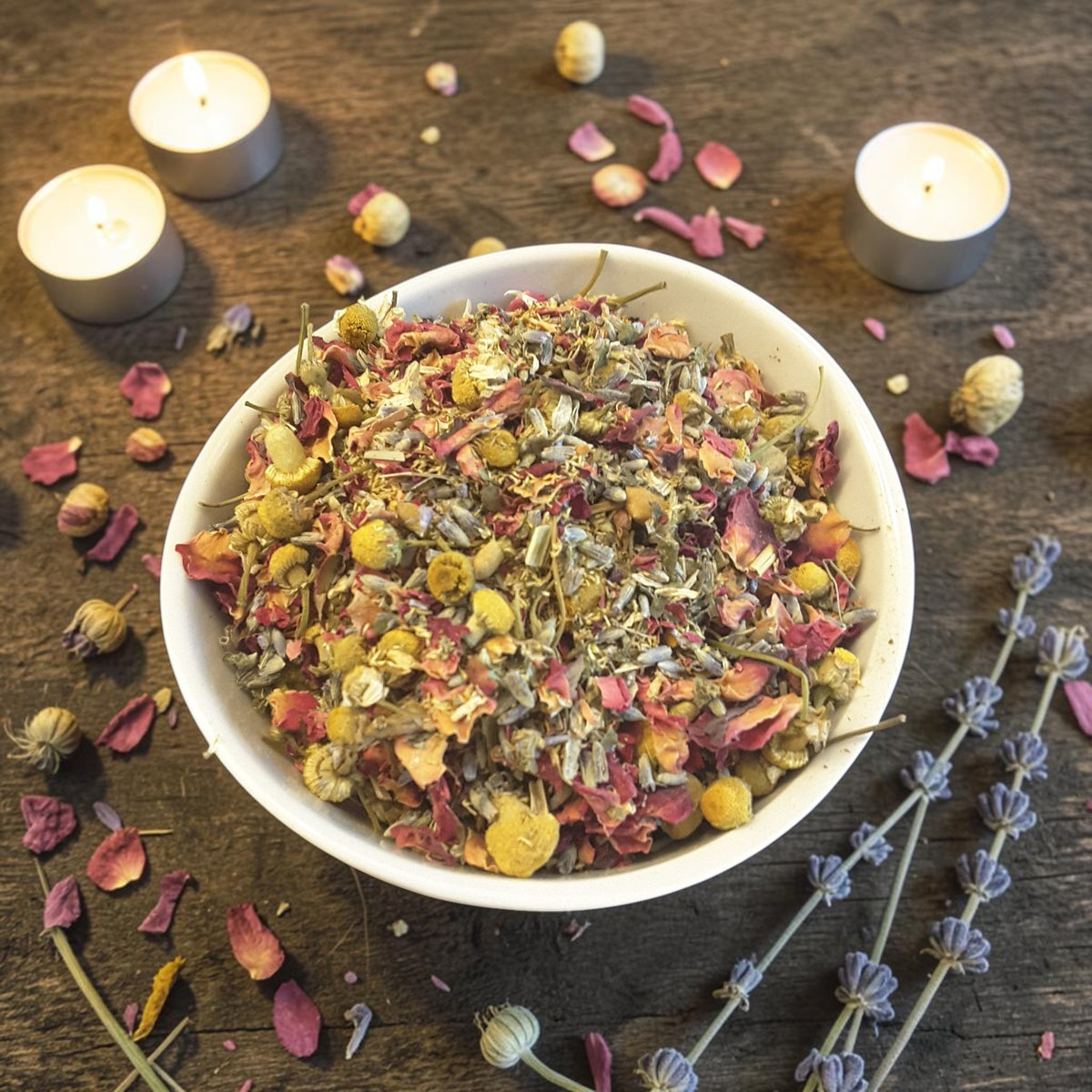 A bowl of peace and calm herbal tub tea  on a wooden surface with candles, lavender and red rose petals