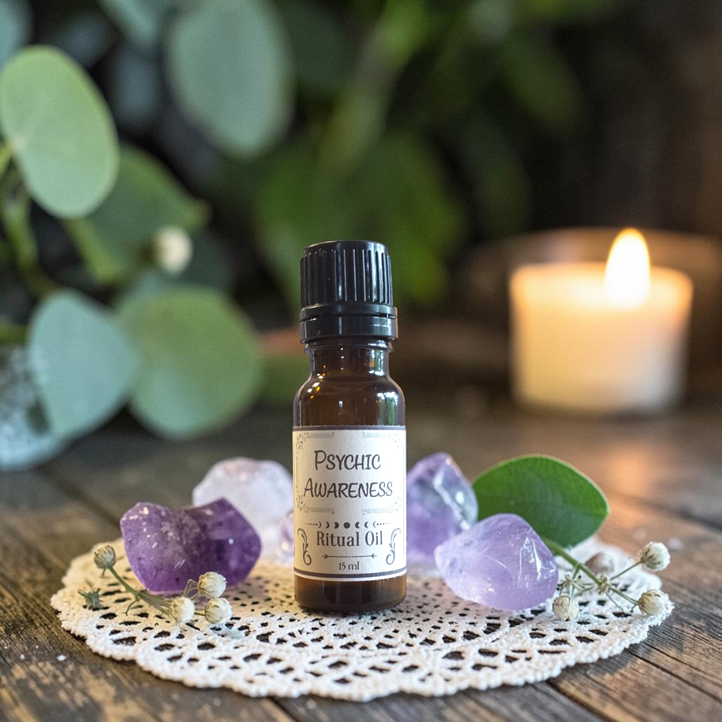 A small amber glass bottle labeled 'Psychic Awareness Ritual Oil' sits on a white lace doily with purple crystals and green leaves, with a blurred lit candle in the background.