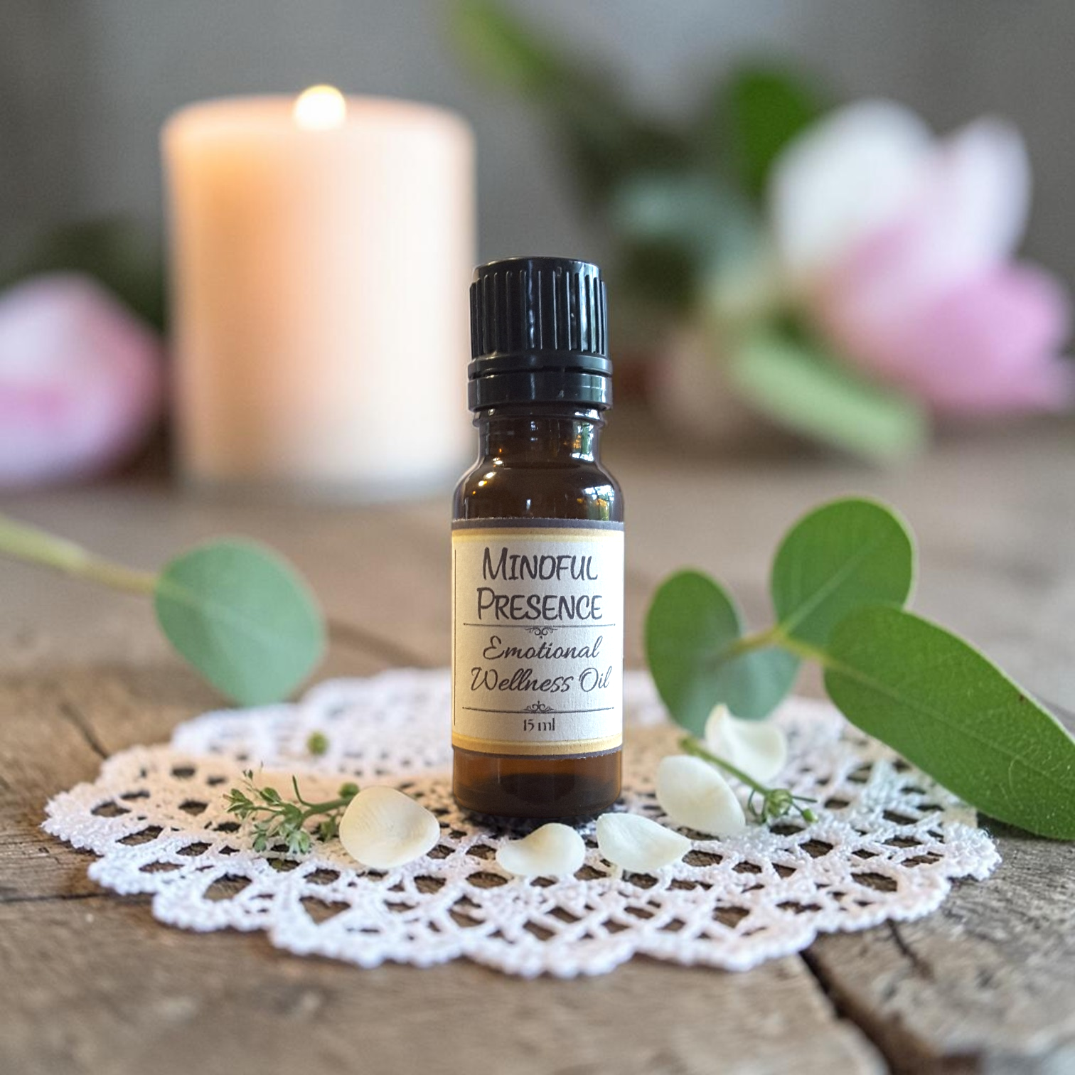 Mindful Presence Emotional Wellness Oil on a doily on a wooden surface with greenery and pink flowers and a candle in the background