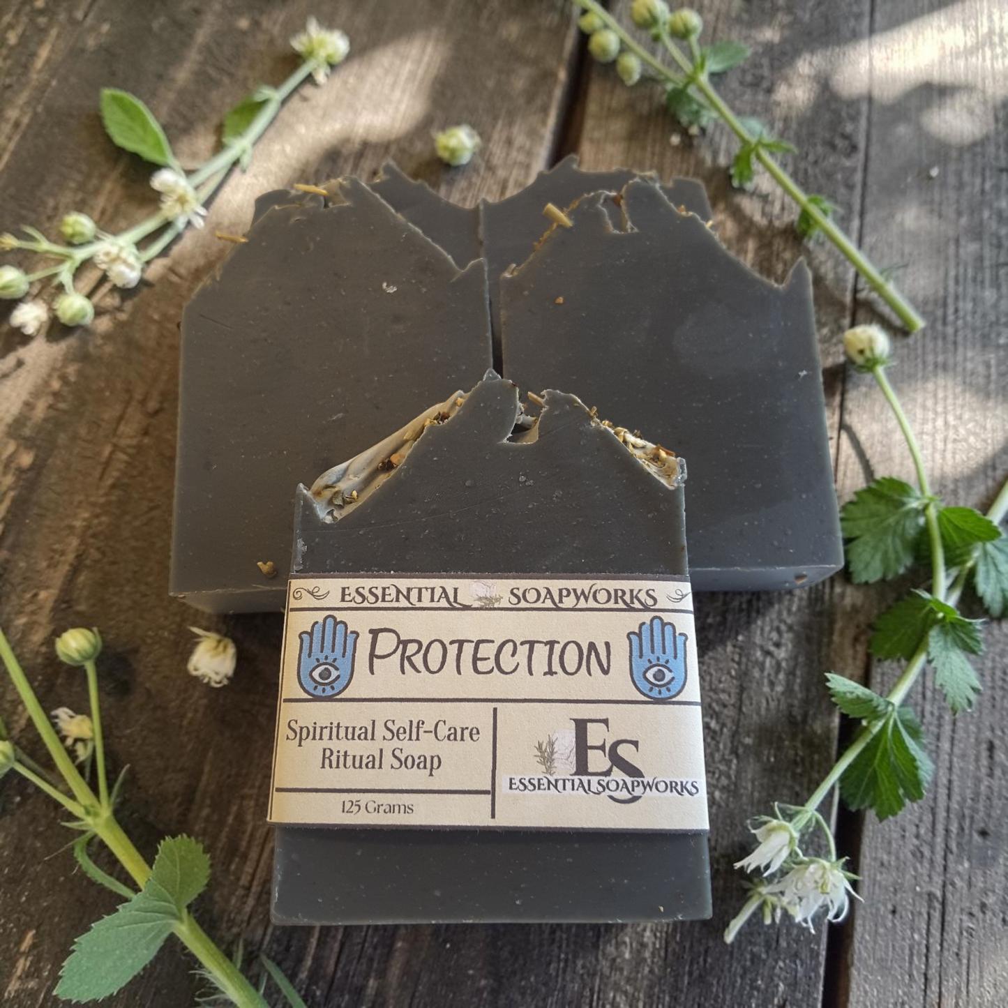 Two dark charcoal gray handmade soap bars with jagged, rustic edges, one wrapped in a paper label that reads 'PROTECTION Spiritual Self-Care Ritual Soap' by 'Essential Soapworks'. The soaps are arranged on a rustic dark wood background decorated with green leaves and small white flowers.