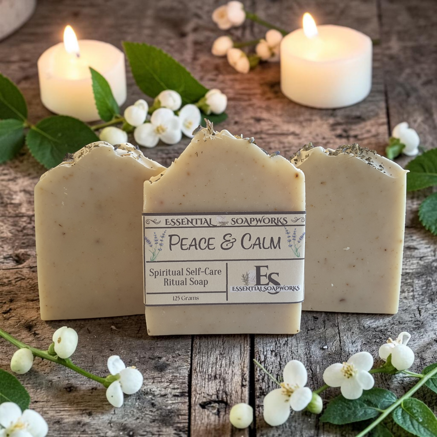 Three bars of Essential Soapworks Peace & Calm ritual soap are arranged on a rustic wooden surface with lit white candles, green leaves, and small white flowers. The center bar is upright and shows the product label.